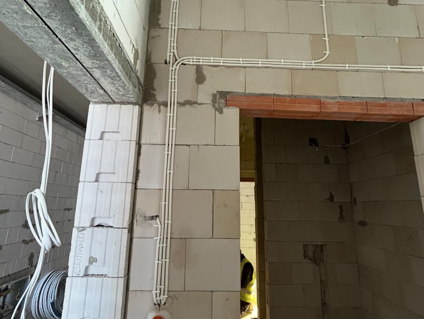 Białe rury instalacji elektrycznej prowadzone po ścianie z bloczków betonu komórkowego w surowym stanie, widoczne kable zwisające z sufitu i fragment pracownika w odblaskowej kamizelce w przejściu.