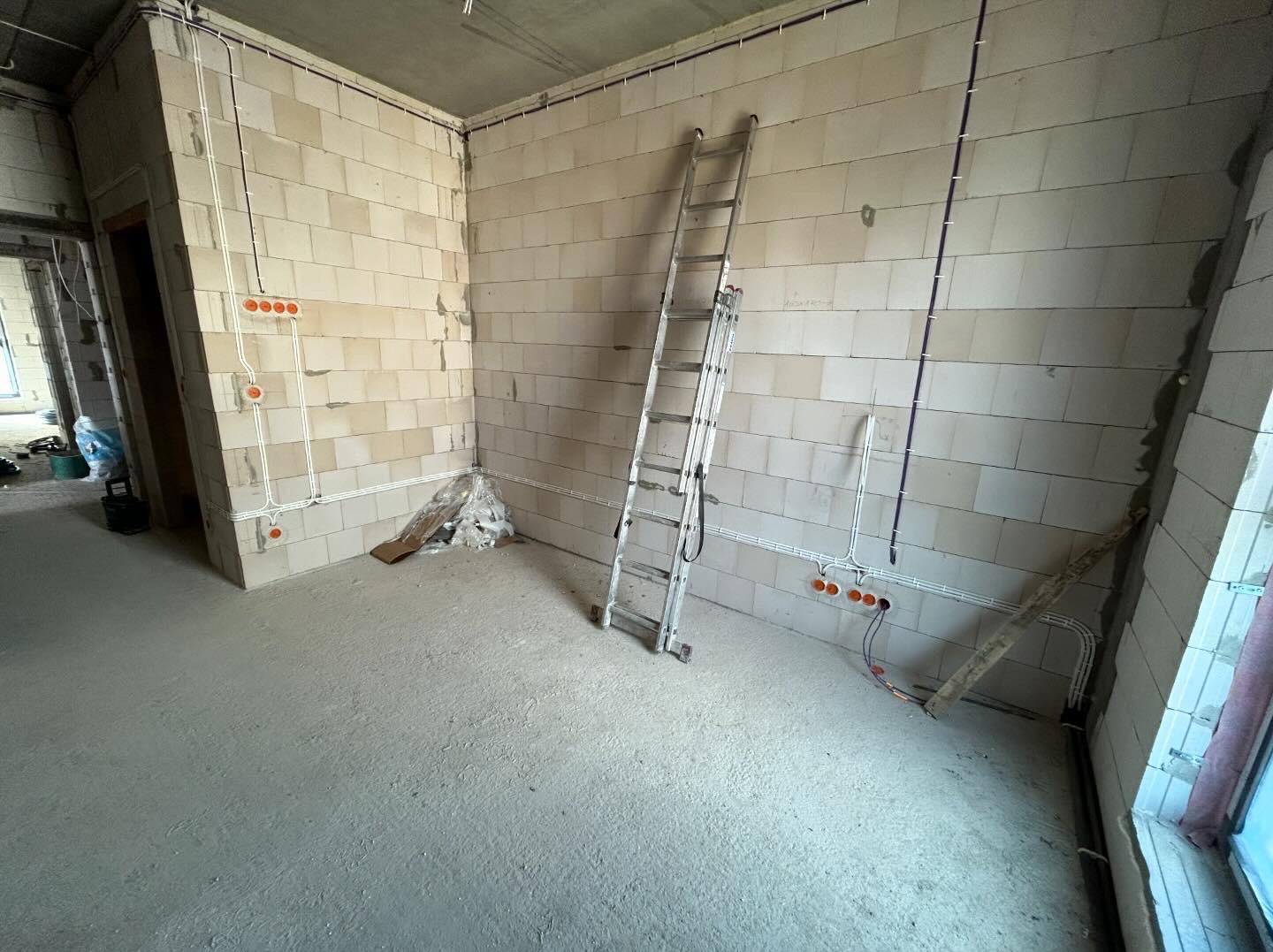 Surowy stan wnętrza z widocznymi przewodami elektrycznymi w białych rurkach na ścianach z betonu komórkowego, aluminiowa drabina oparta o ścianę, betonowa wylewka na podłodze.