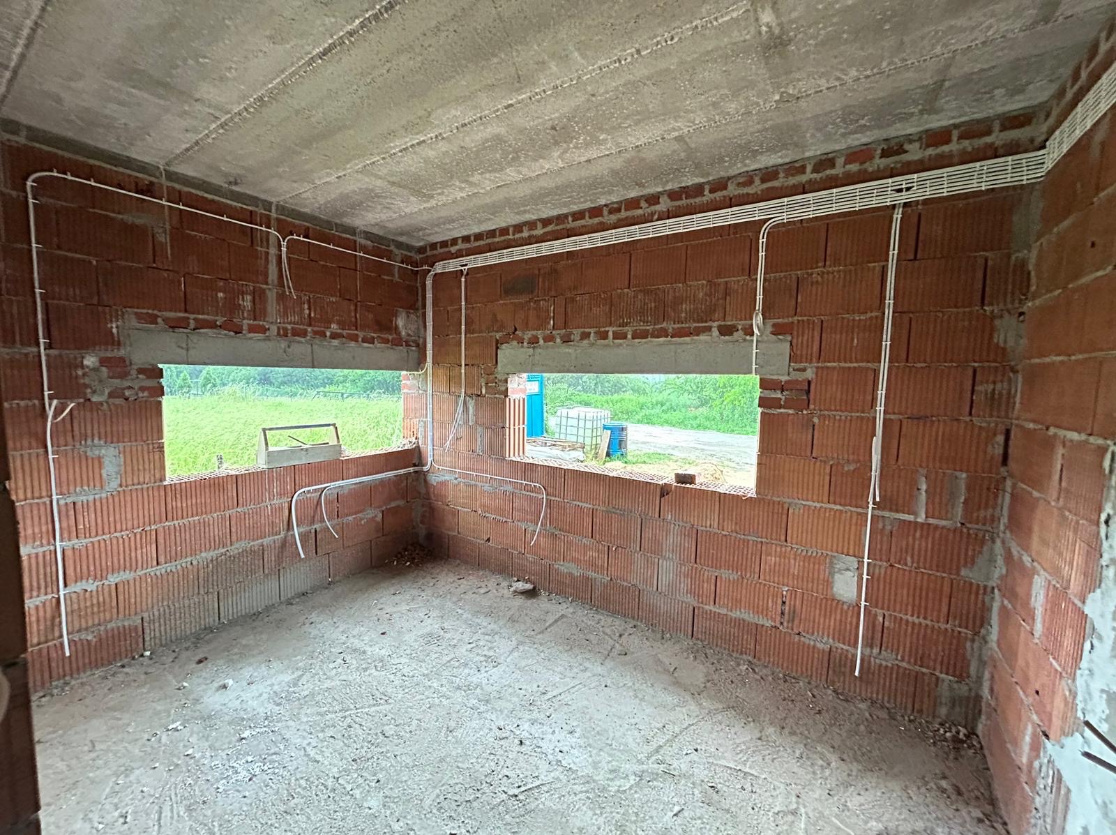 Wnętrze surowego budynku z widoczną instalacją elektryczną w białych listwach, prowadzona po ścianach z czerwonej cegły i pod betonowym sufitem, okna z widokiem na zieleń.