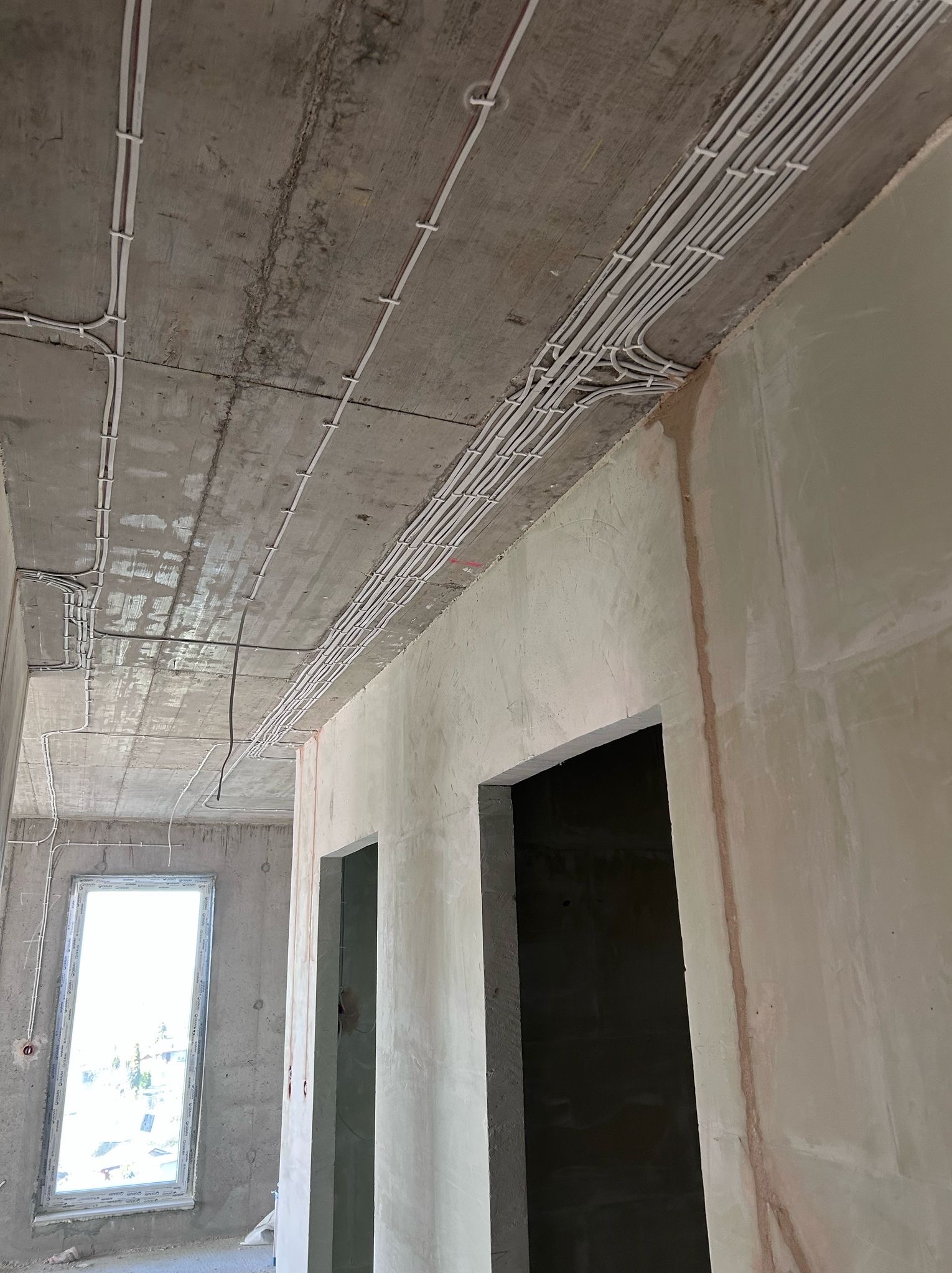 Białe rurki instalacji elektrycznej prowadzone po surowym, betonowym suficie w nowym budynku, widoczne otwory drzwiowe i okienne, ściany w stanie surowym.