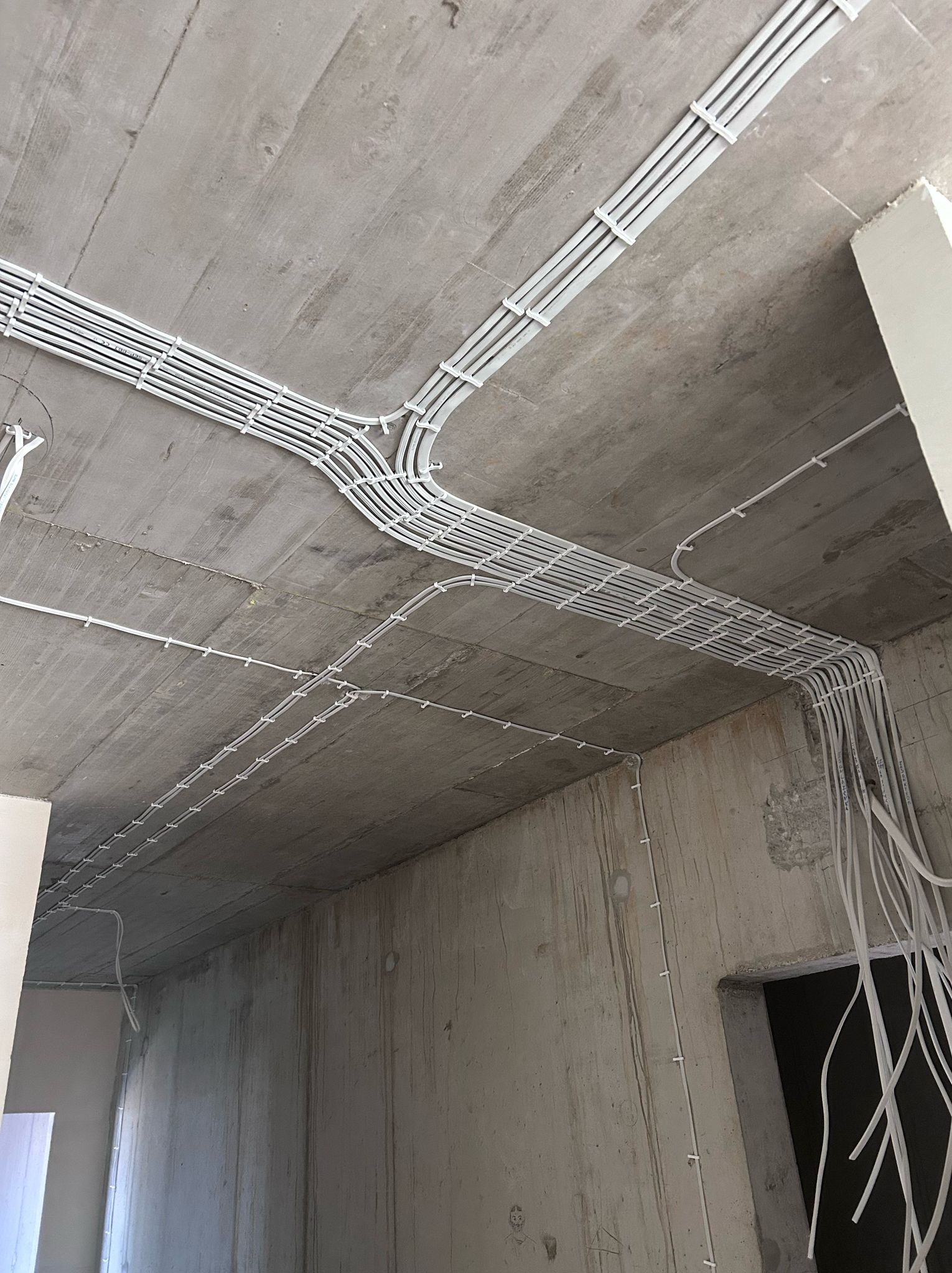 Montaż instalacji elektrycznej w surowym wnętrzu, widoczne białe korytka i przewody na betonowym suficie i ścianach, etap prac wykończeniowych.