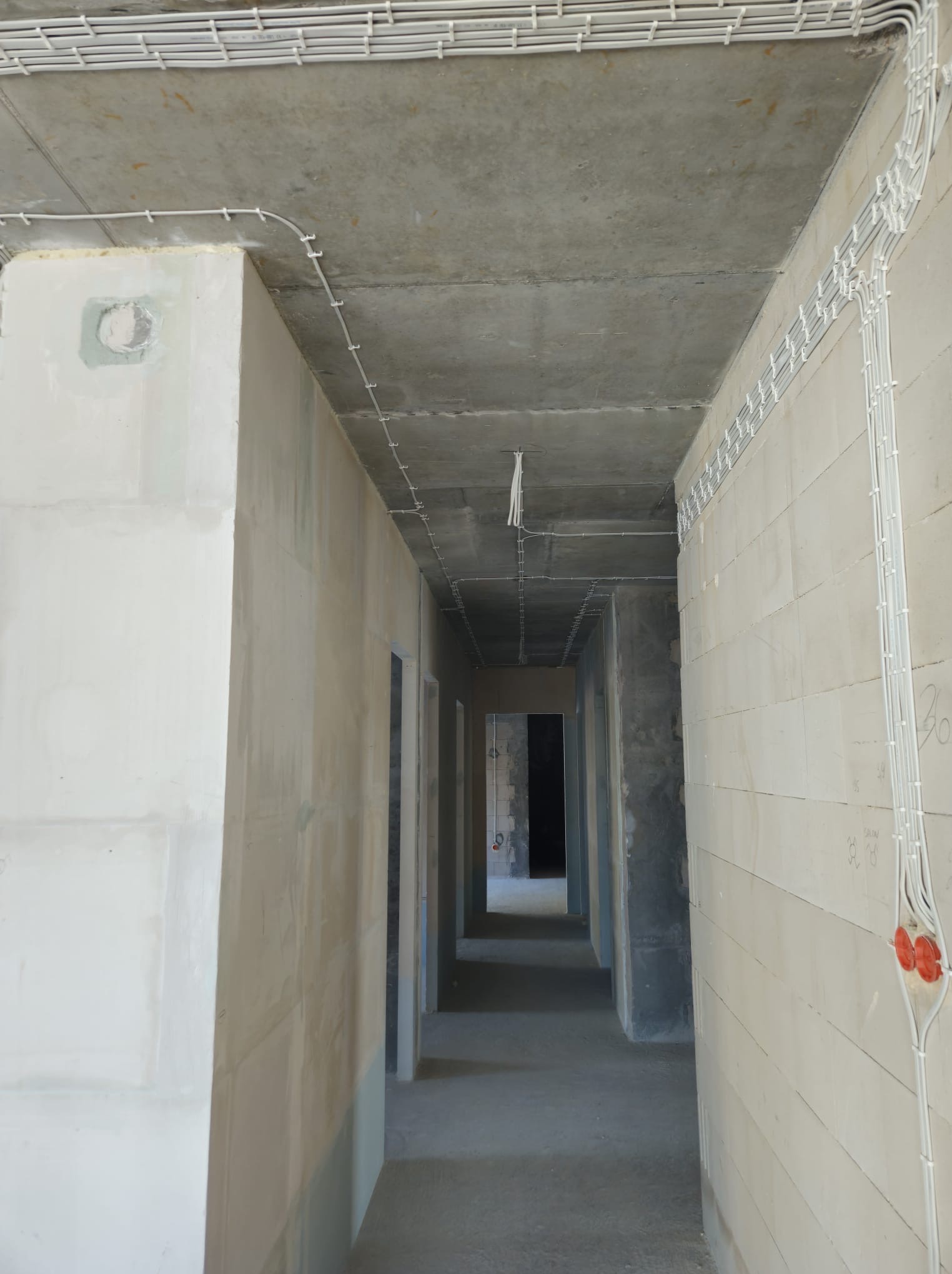 Korytarz w stanie surowym z widoczną instalacją kablową pod sufitem i na ścianach, uchwyty kablowe w równych odstępach, puszki elektryczne w kolorze czerwonym na ścianie z betonu komórkowego.