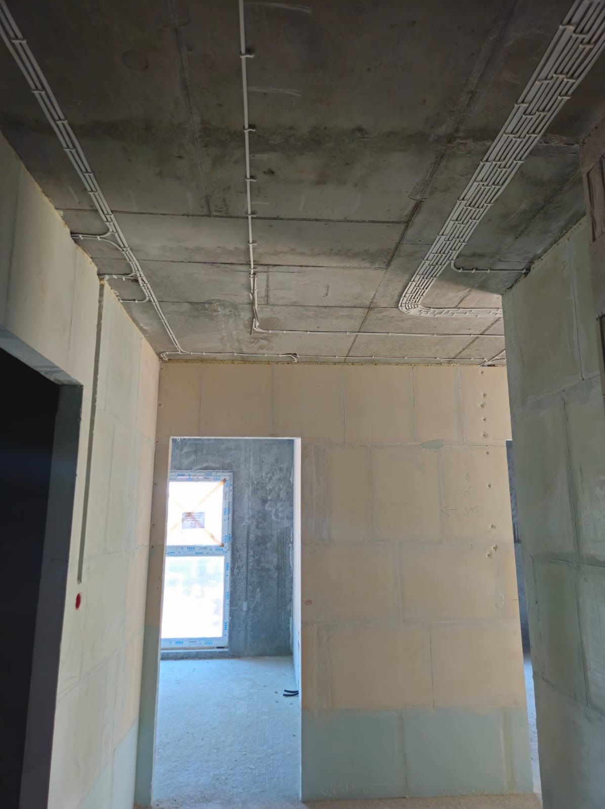 Kable elektryczne prowadzone po suficie w nowym budynku w stanie deweloperskim, widoczne białe korytka kablowe i przewody, surowy betonowy strop i ściany z bloczków.