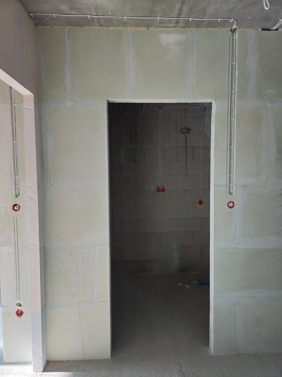 Surowy stan wnętrza w trakcie prac, widoczne kable elektryczne wyprowadzone ze ściany i sufitu, przygotowane otwory na gniazdka, ściany z białych bloczków.