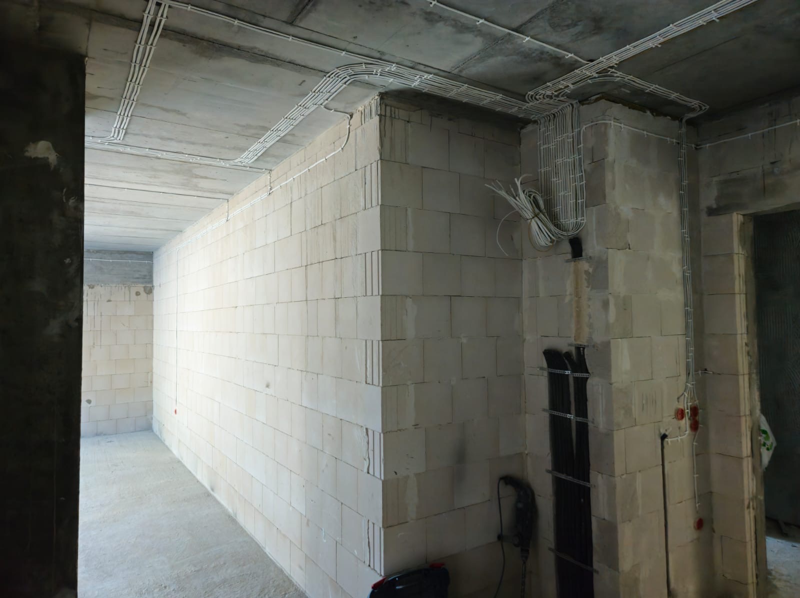 Wnętrze w stanie surowym z widoczną instalacją elektryczną w białych rurkach na suficie i ścianach z bloczków. Część przewodów zwisa ze ściany. Widoczne narzędzia na podłodze.