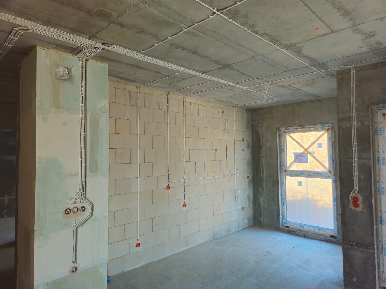 Surowy stan wnętrza z widoczną instalacją elektryczną: kable w białych listwach prowadzone po suficie i ścianach z betonu komórkowego, przygotowane punkty pod gniazdka i włączniki, okno...