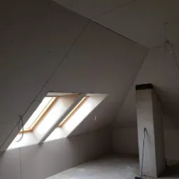 Poddasze po montażu płyt gipsowo-kartonowych, widoczne dwa okna dachowe w drewnianych ramach, komin i częściowo posprzątana podłoga.