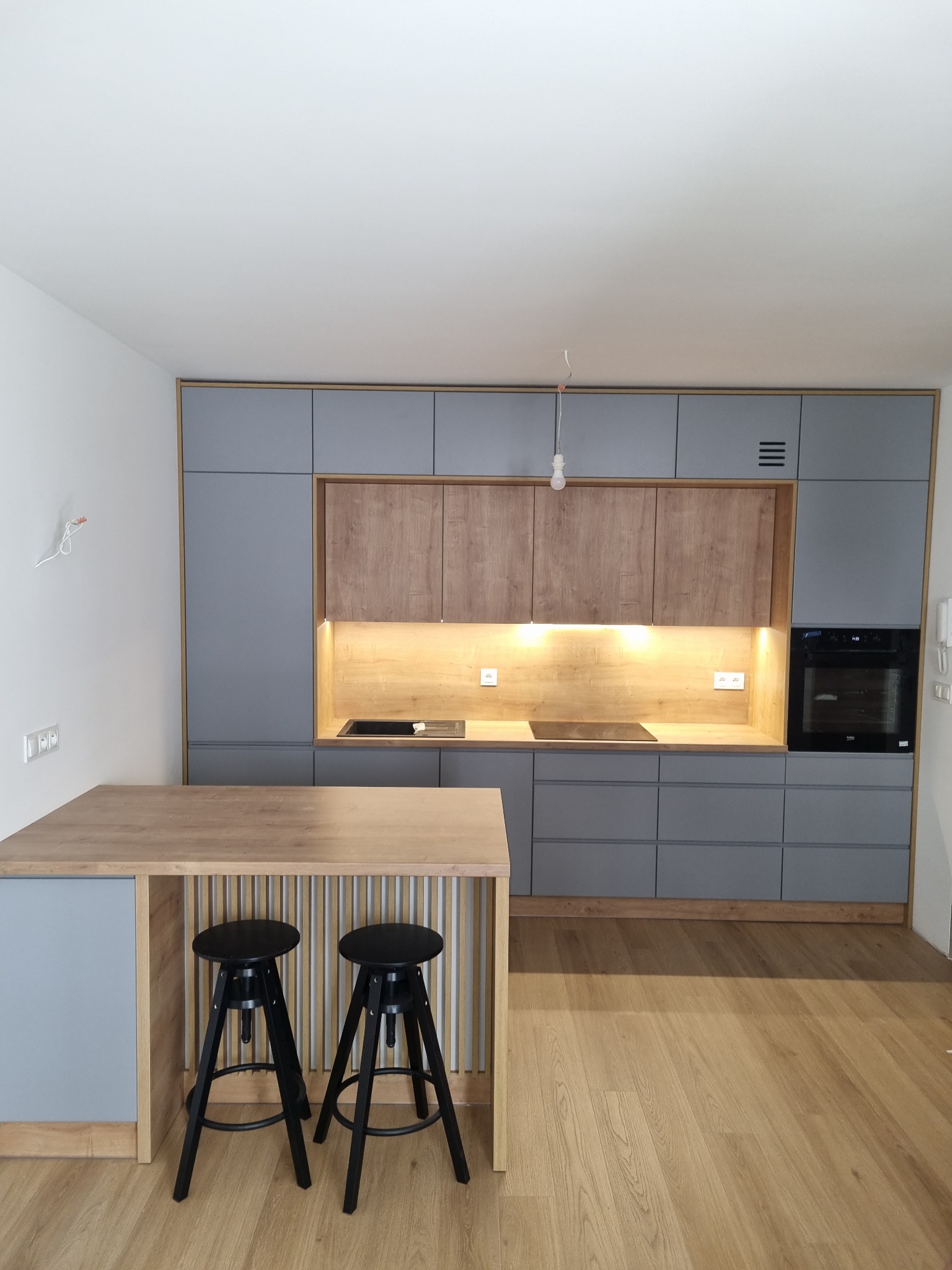 Nowoczesna kuchnia z szarymi szafkami i drewnianymi akcentami, podświetlane blaty, czarne hokery przy drewnianym blacie, minimalistyczny design.