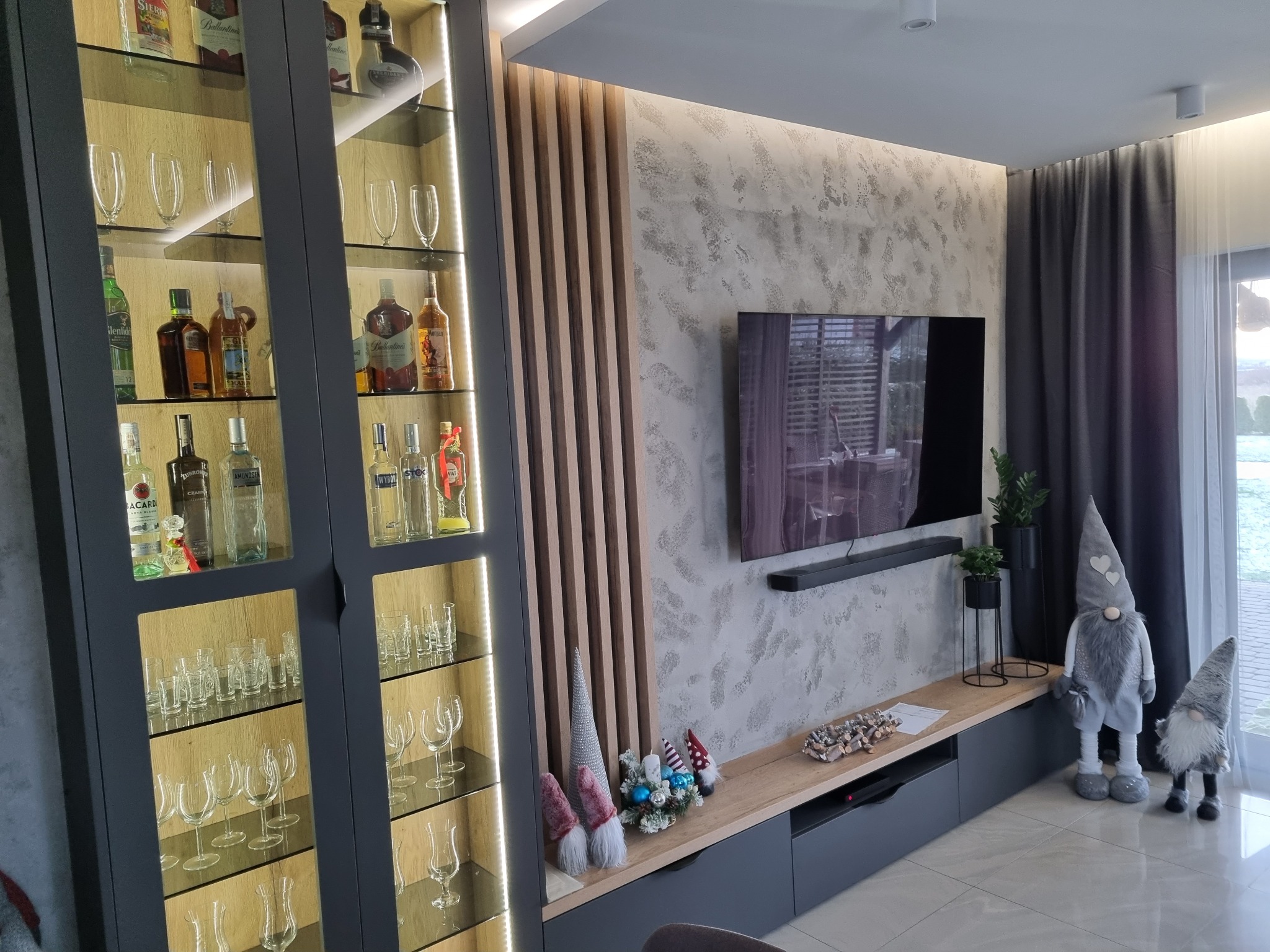 Nowoczesny salon z podświetlaną witryną wypełnioną szkłem i alkoholem, drewnianymi lamelami na ścianie oraz telewizorem na tle ściany o nieregularnej fakturze.