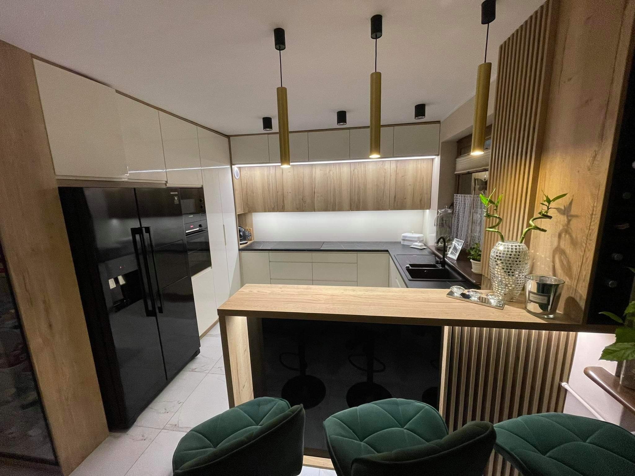 Nowoczesna kuchnia z wyspą i hokerami w kolorze butelkowej zieleni, oświetlenie punktowe i wiszące lampy tuby, drewniane akcenty na meblach i ścianie.