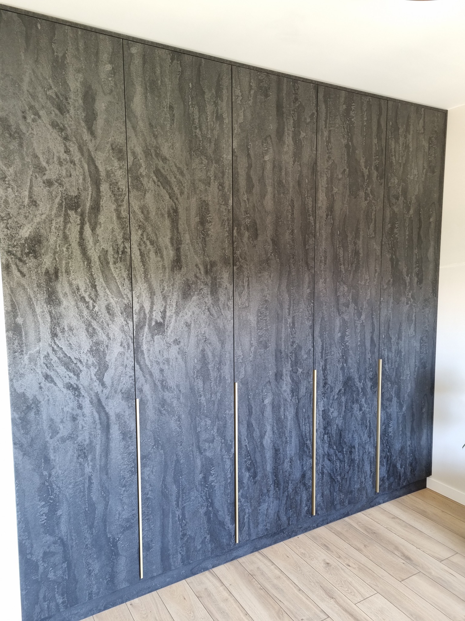 Duża szafa wnękowa z frontami imitującymi kamień, z długimi, pionowymi, złotymi uchwytami, na tle jasnej podłogi z paneli.