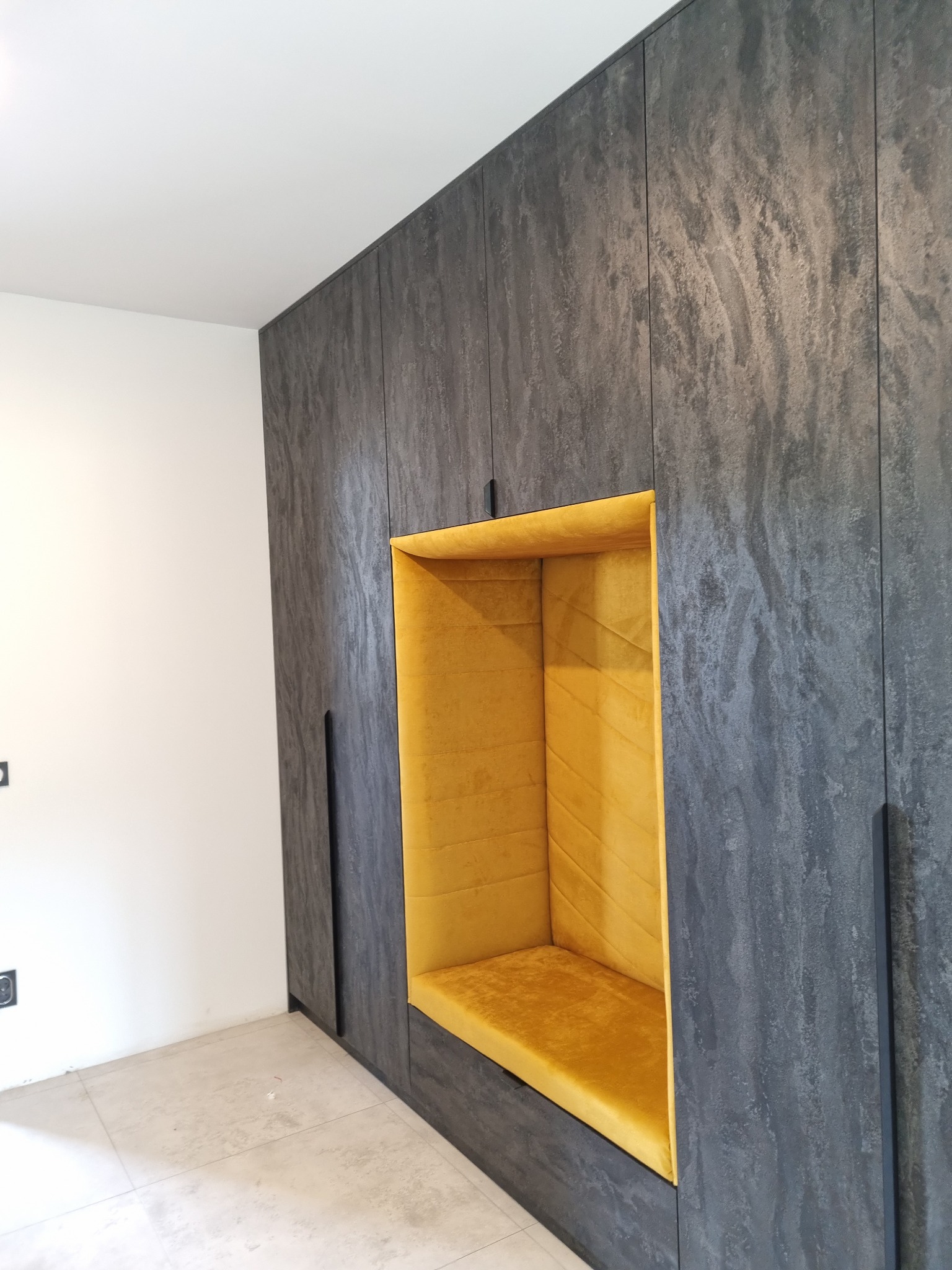 Wnęka z siedziskiem obita żółtą tkaniną, wkomponowana w zabudowę ścienną o szarym, kamiennym wykończeniu, z czarnymi, minimalistycznymi uchwytami.