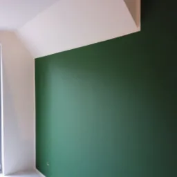 Świeżo pomalowane wnętrze z zieloną ścianą i białym sufitem pod skosem, widoczny fragment okna i surowa podłoga przed wykończeniem.