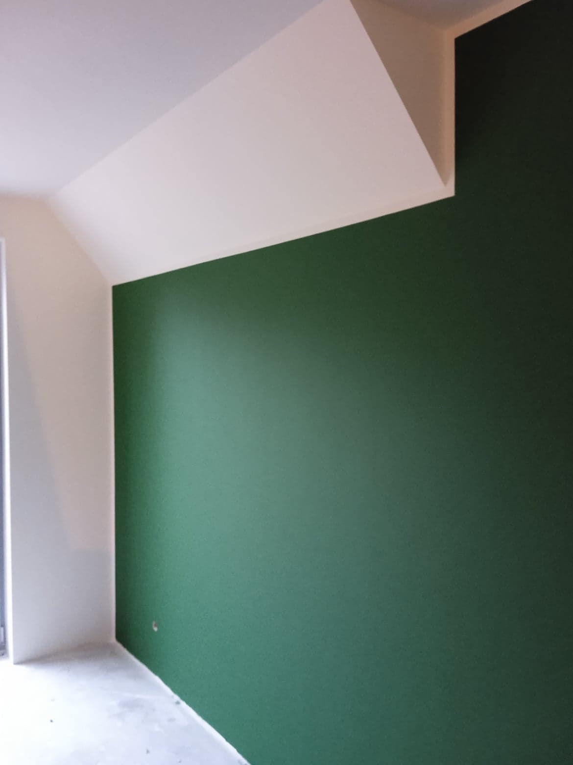 Świeżo pomalowane wnętrze z zieloną ścianą i białym sufitem pod skosem, widoczny fragment okna i surowa podłoga przed wykończeniem.