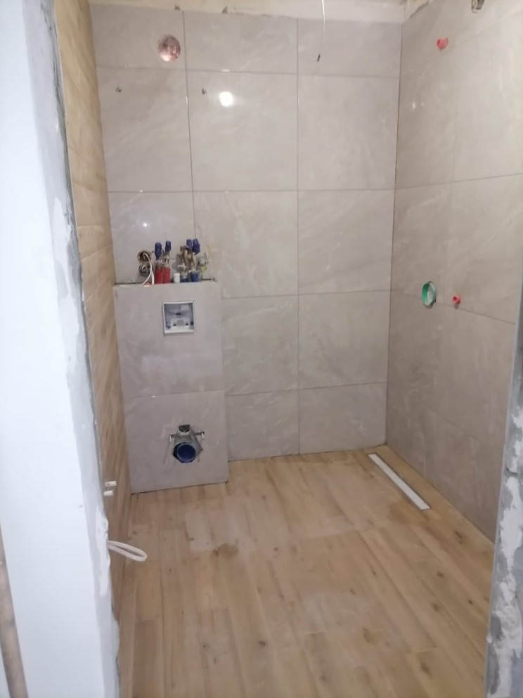 Wnętrze łazienki w trakcie remontu z położonymi płytkami imitującymi drewno na podłodze i jasnymi płytkami na ścianach, widoczne elementy instalacji wodnej i kanalizacyjnej, surowy stan wykończenia...