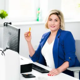 Kobieta w niebieskiej marynarce siedzi przy biurku z komputerem, trzymając żółty długopis i uśmiechając się.