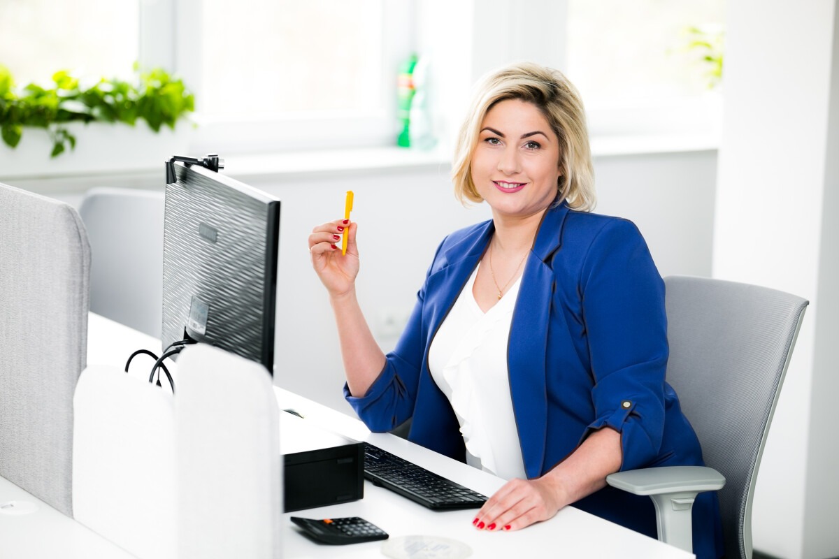 Kobieta w niebieskiej marynarce siedzi przy biurku z komputerem, trzymając żółty długopis i uśmiechając się.