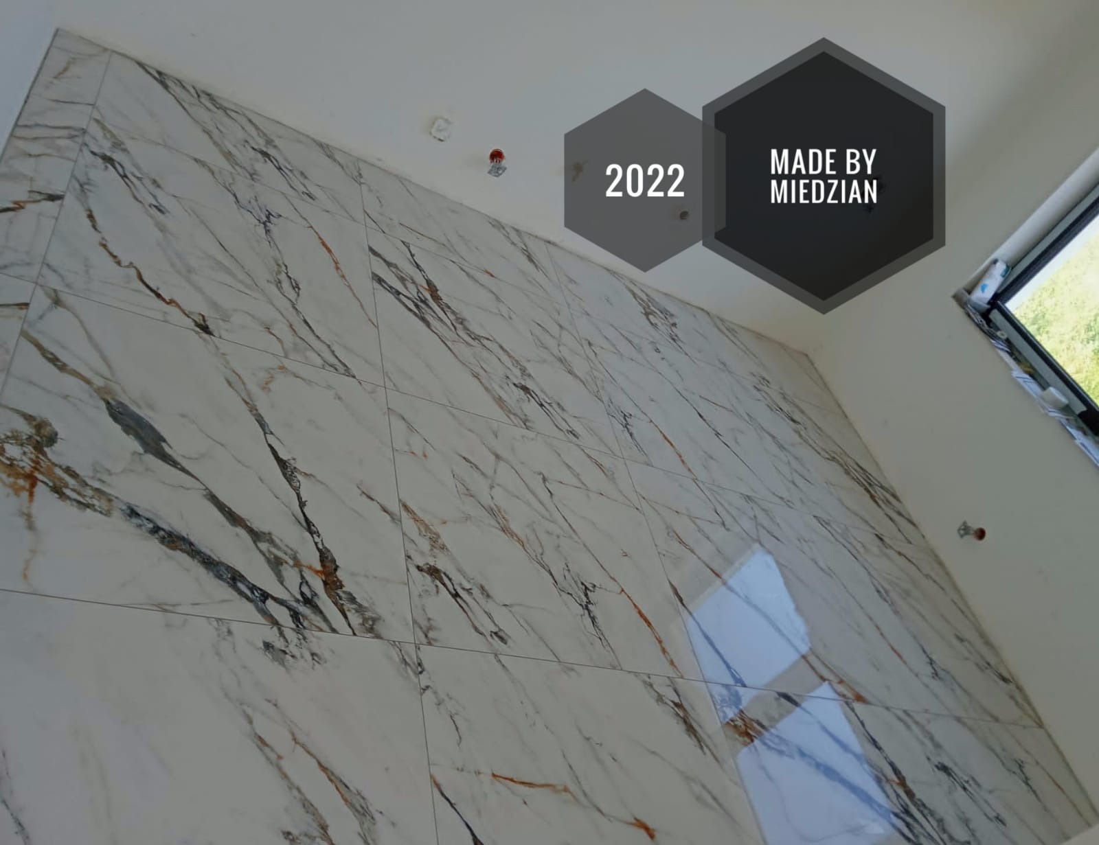 Wykończona ściana z płytek imitujących marmur z ciemnymi żyłami, widoczne logo firmy 'Made by Miedzian' z rokiem 2022 w górnym rogu
