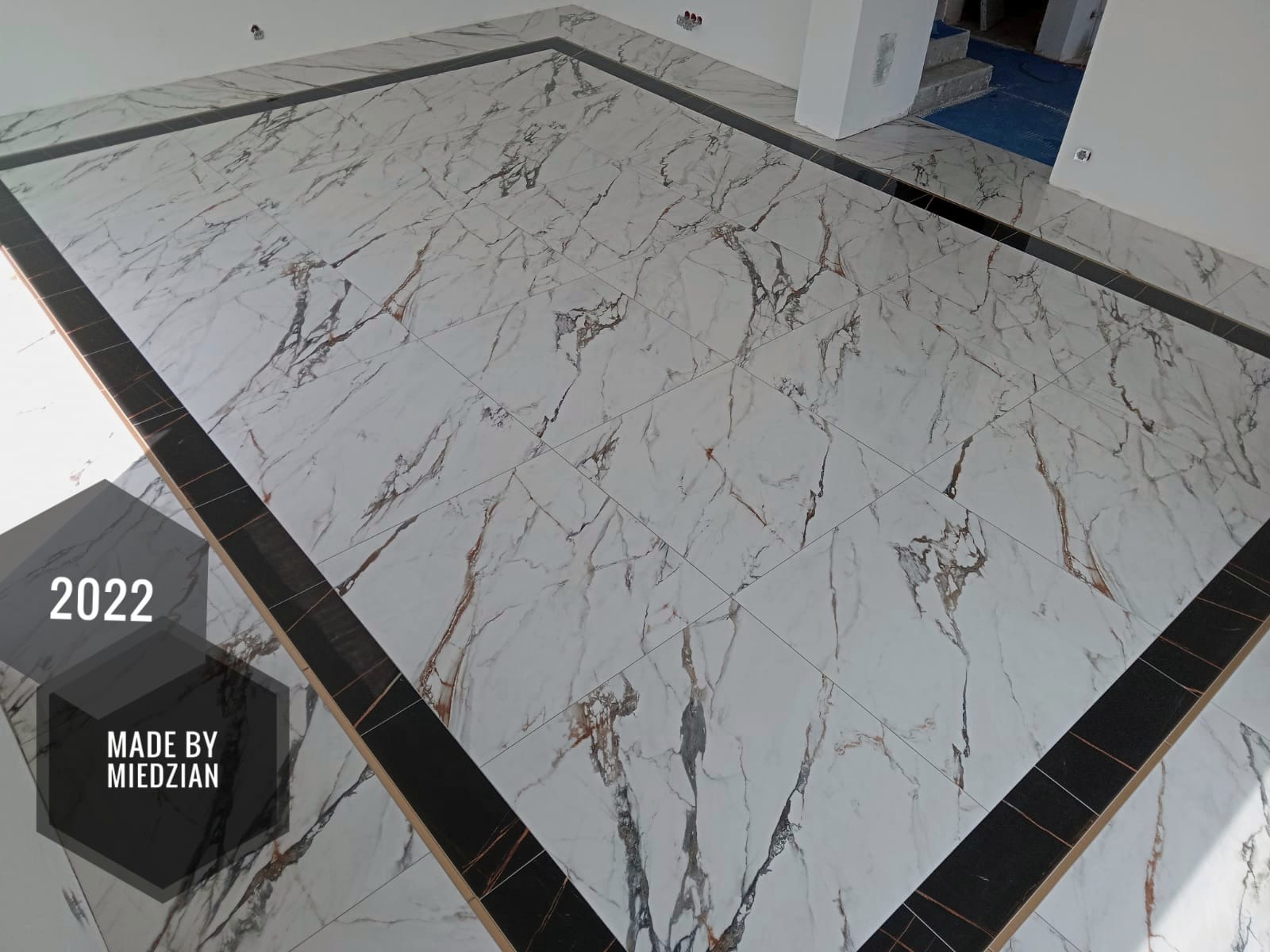 Wykończona podłoga z białych płytek imitujących marmur z czarną ramką, widoczny fragment logo firmy wykonującej.