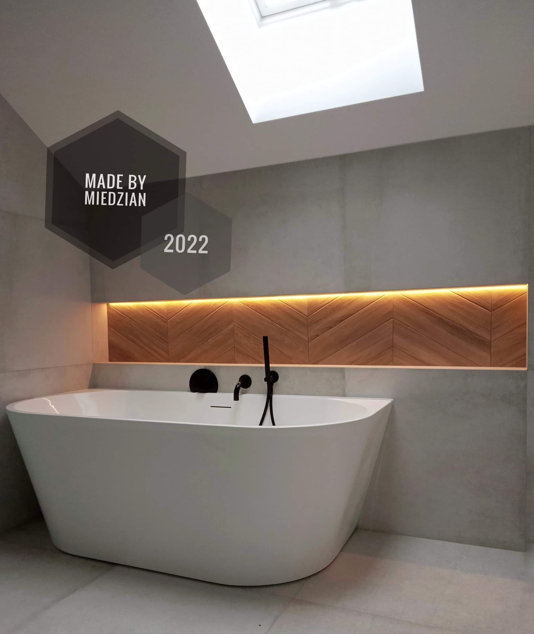 Nowoczesna łazienka z wolnostojącą białą wanną, podświetlaną wnęką z drewnopodobną mozaiką ułożoną we wzór jodełki, szare płytki na ścianach i podłodze, oraz oknem dachowym. Na zdjęciu widoczny...