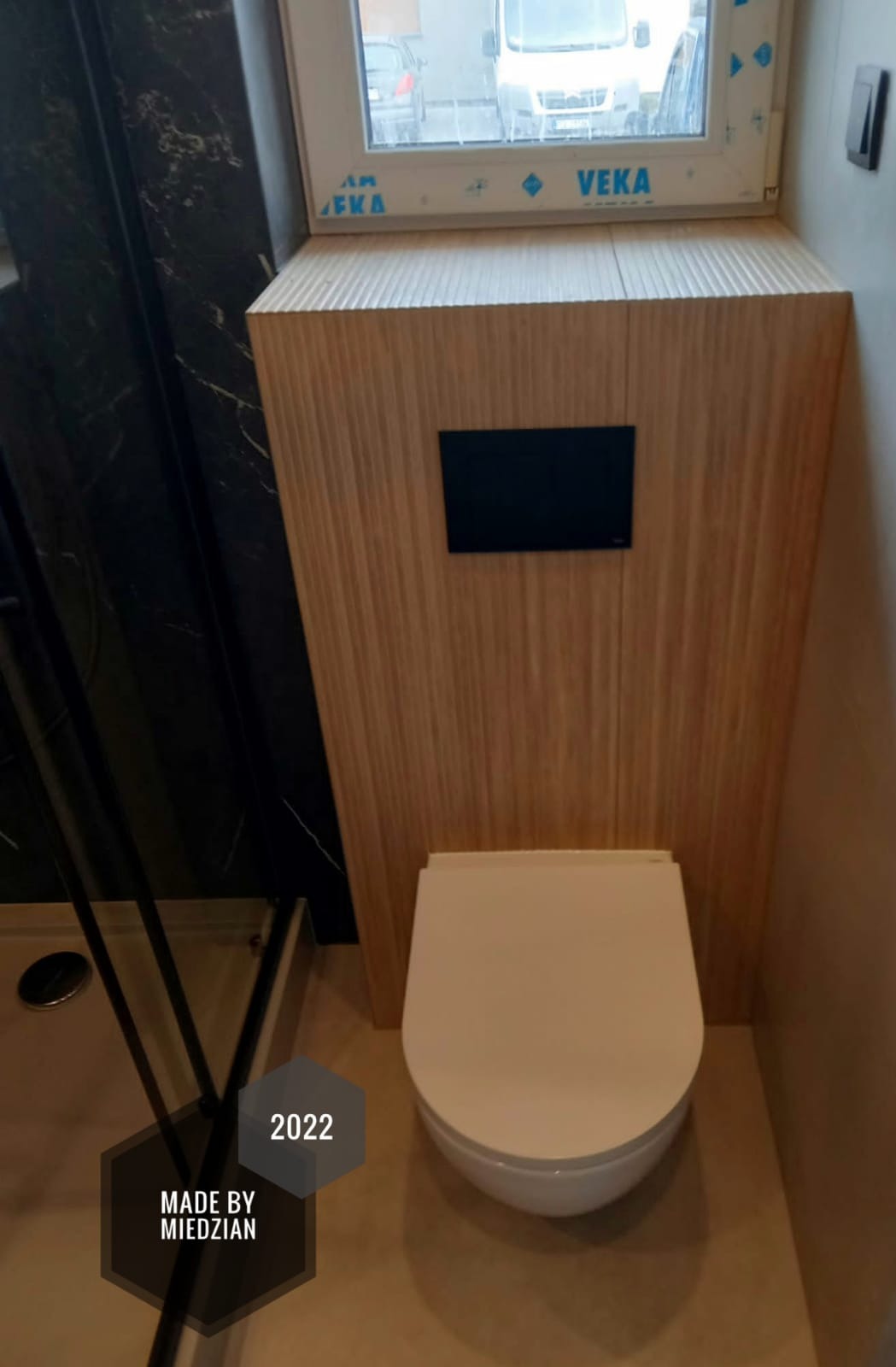 Wisząca toaleta z zabudową z paneli drewnopodobnych, czarny panel sterujący spłukiwaniem, fragment kabiny prysznicowej z czarnym wykończeniem, okno z widokiem na samochody.