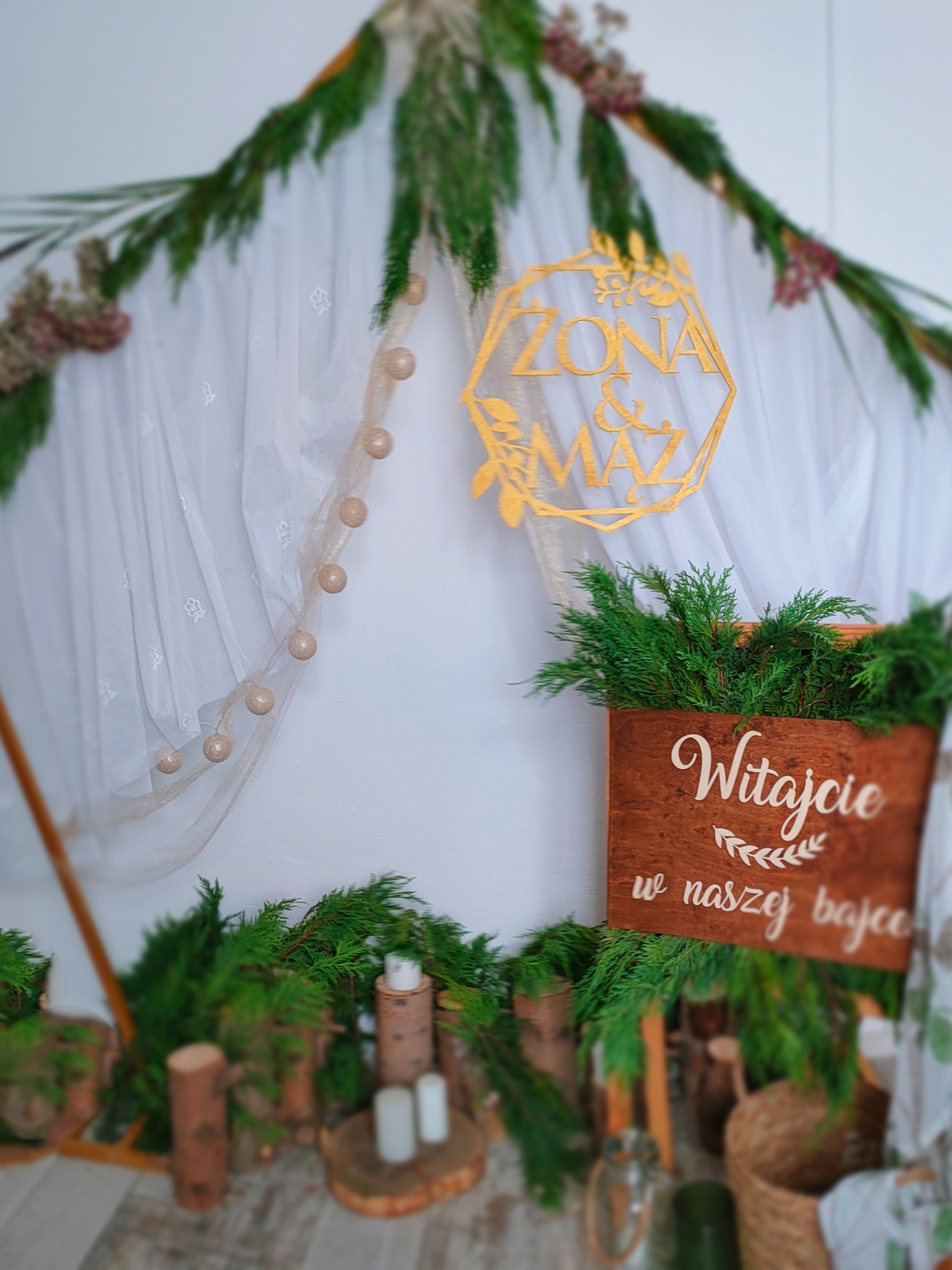 Dekoracja ślubna w stylu rustykalnym: drewniane elementy, zieleń, białe tkaniny, napis 'Żona & Mąż' oraz 'Witajcie w naszej bajce'.