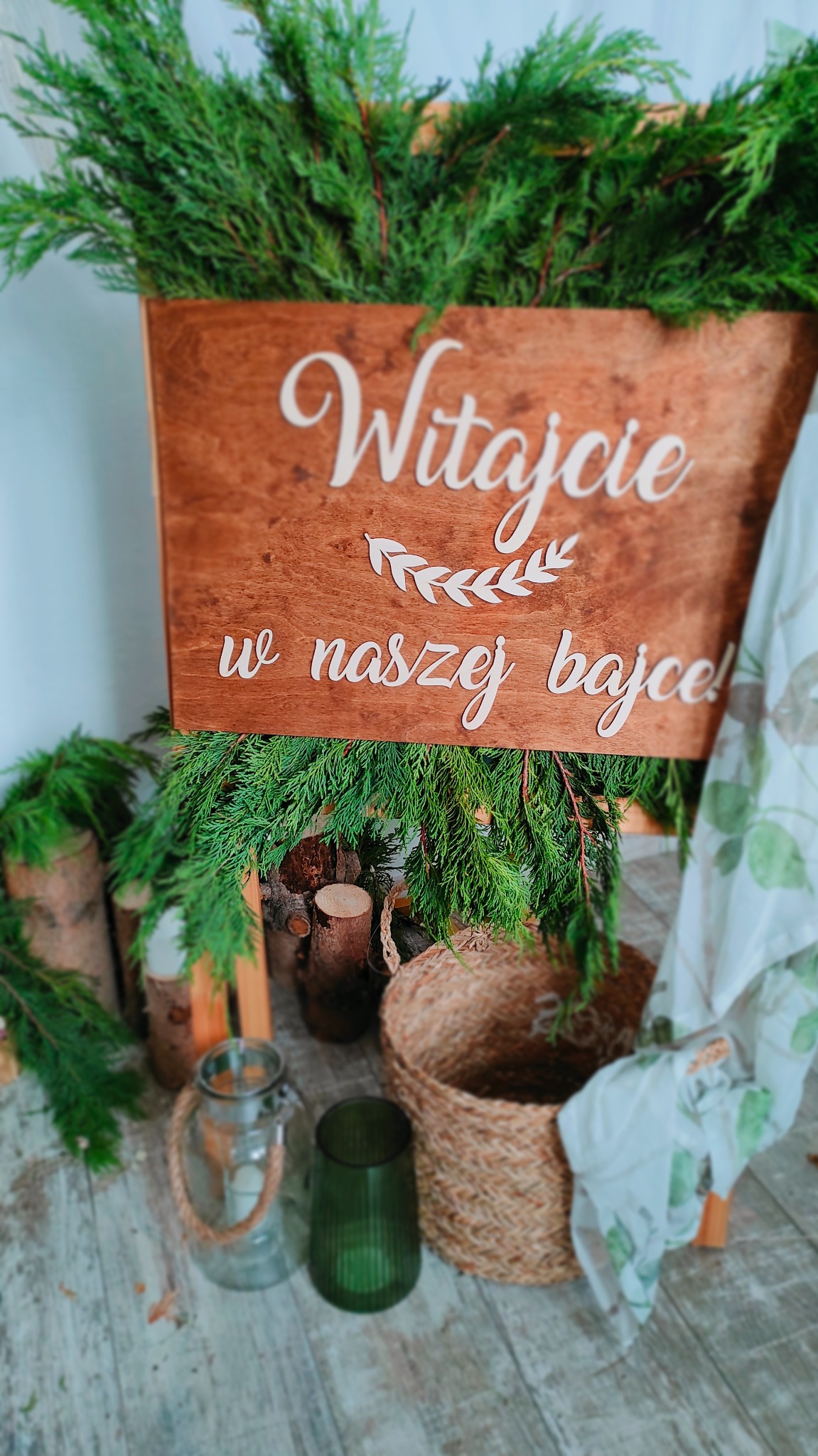 Drewniana tablica powitalna z napisem 'Witajcie w naszej bajce!' otoczona zielonymi gałązkami, drewnianymi pieńkami, szklanymi lampionami i wiklinowym koszem, tworząca rustykalną dekorację.