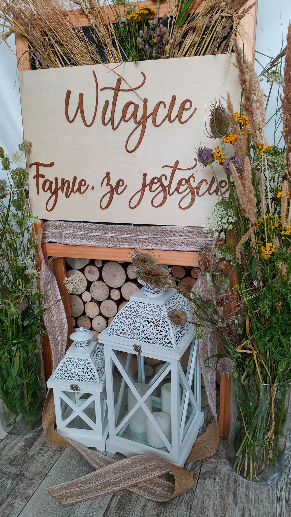 Dekoracja rustykalna na powitanie gości: drewniana tablica z napisem 'Witajcie. Fajnie, że jesteście', otoczona kompozycjami z polnych kwiatów i traw, z białymi lampionami i pniem drewna w tle.