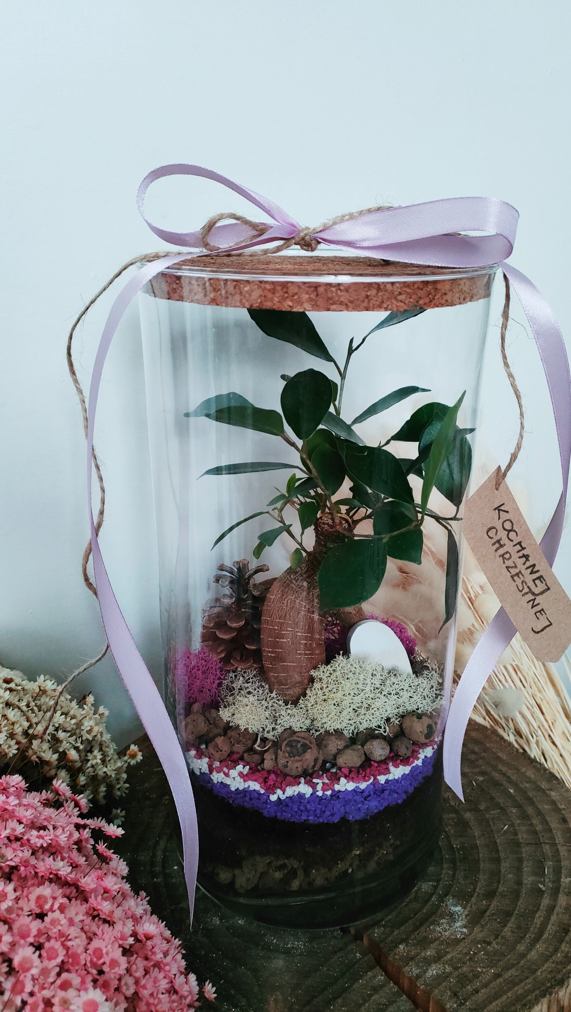 Szklany słój z miniaturowym ekosystemem: bonsai, mech, szyszka, warstwy kolorowego żwirku i wstążka z napisem 'Kochanej Chrzestnej'.
