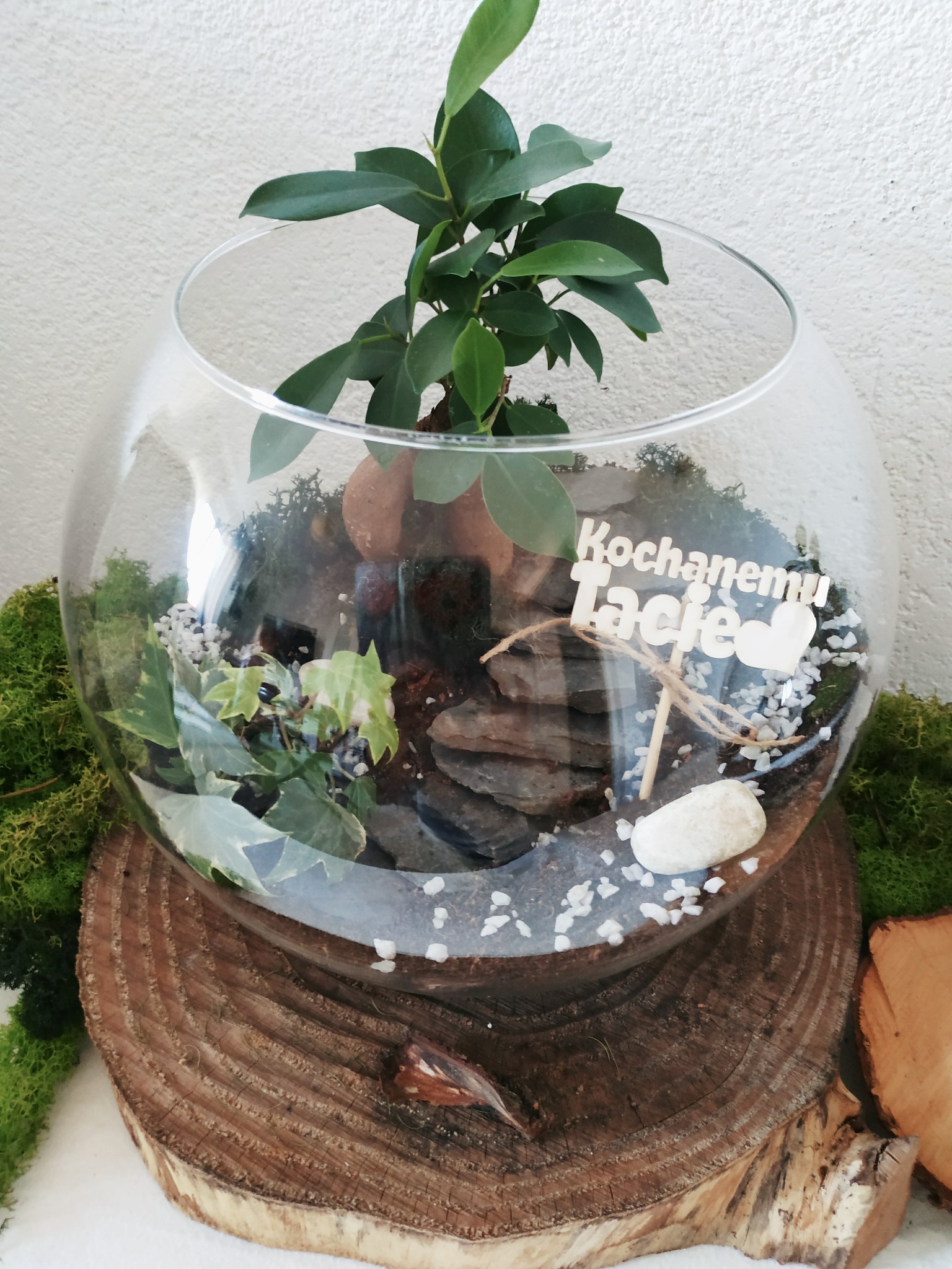 Szklane terrarium z roślinami, kamieniami i dekoracją 'Kochanemu Tacie', umieszczone na drewnianym postumencie z mchem.
