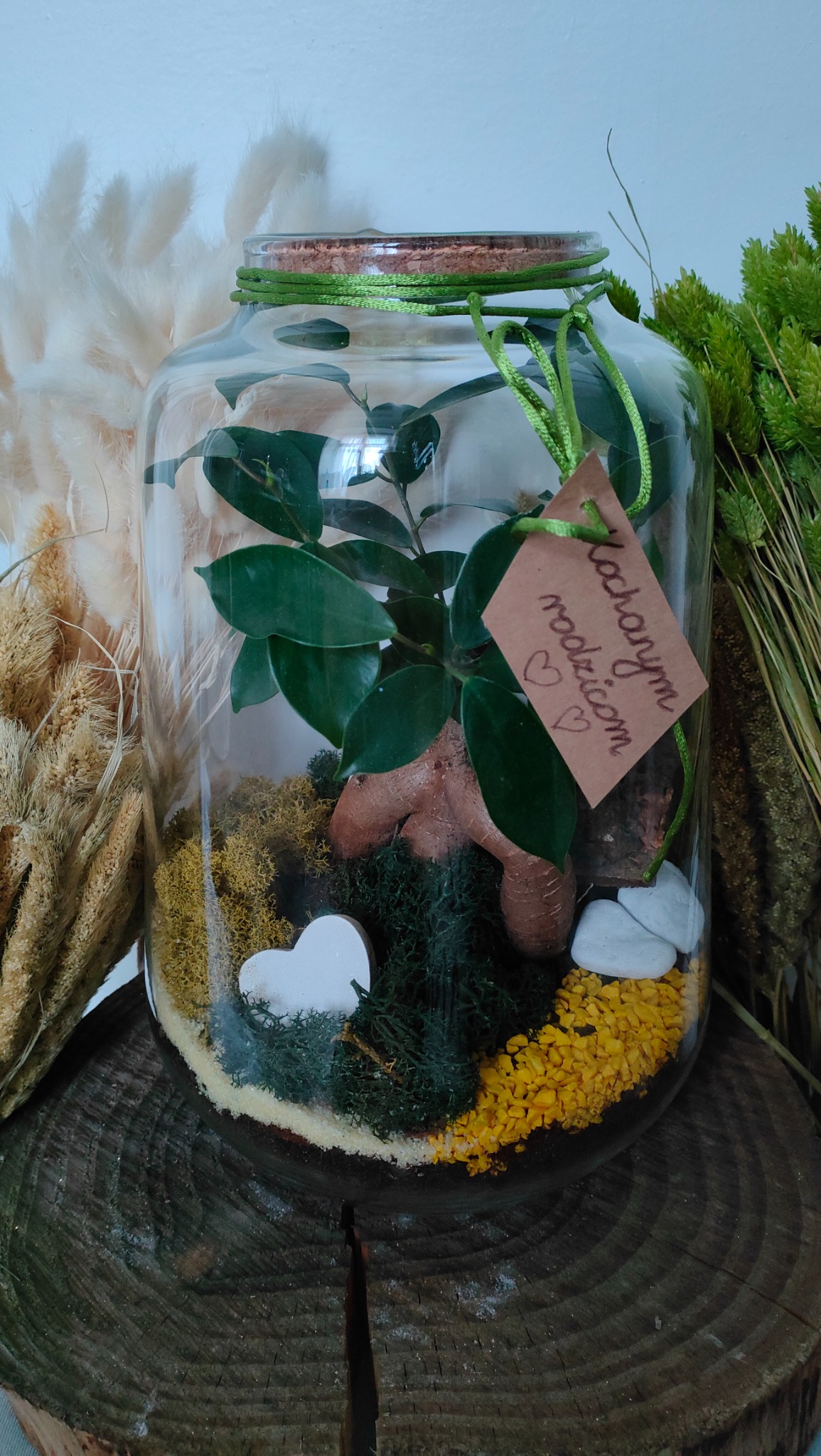Szklany słój z kompozycją roślinną, mchem, kamykami i drewnianym sercem, przewiązany sznurkiem, z zawieszką z napisem 'Kochanym Rodzicom' na tle suszonych traw, prezentowany przez florystę z Bielawy.