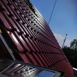 Czerwona dachówka ceramiczna ułożona na skośnym dachu z widocznym oknem dachowym i fragmentem nieba w tle.