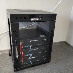 Czarna szafa rack z trzema modułami bateryjnymi Heckman RLFPS1100A, podłączonymi przewodami z czerwonymi i czarnymi końcówkami oraz niebieskimi kablami Ethernet, umieszczona na szarym dywanie...