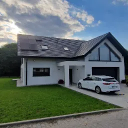 Nowoczesny dom jednorodzinny z panelami słonecznymi na dachu, widok z zewnątrz, biały samochód zaparkowany na podjeździe.