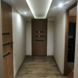 Korytarz z drewnopodobną podłogą, białymi ścianami i oświetleniem sufitowym LED, widoczne trzy drewniane drzwi z przeszkleniami.