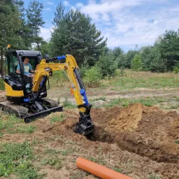 Żółto-czarna mini koparka SANY z operatorem w kabinie, wykonuje wykop w ziemi na działce z drzewami w tle; obok wykopu leży pomarańczowa rura.