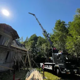 Czarny dźwig z wysięgnikiem podnosi elementy więźby dachowej na budowę domu jednorodzinnego w słoneczny dzień. Na pierwszym planie stos kantówek. Na dachu pracownik w trakcie układania dachówki.