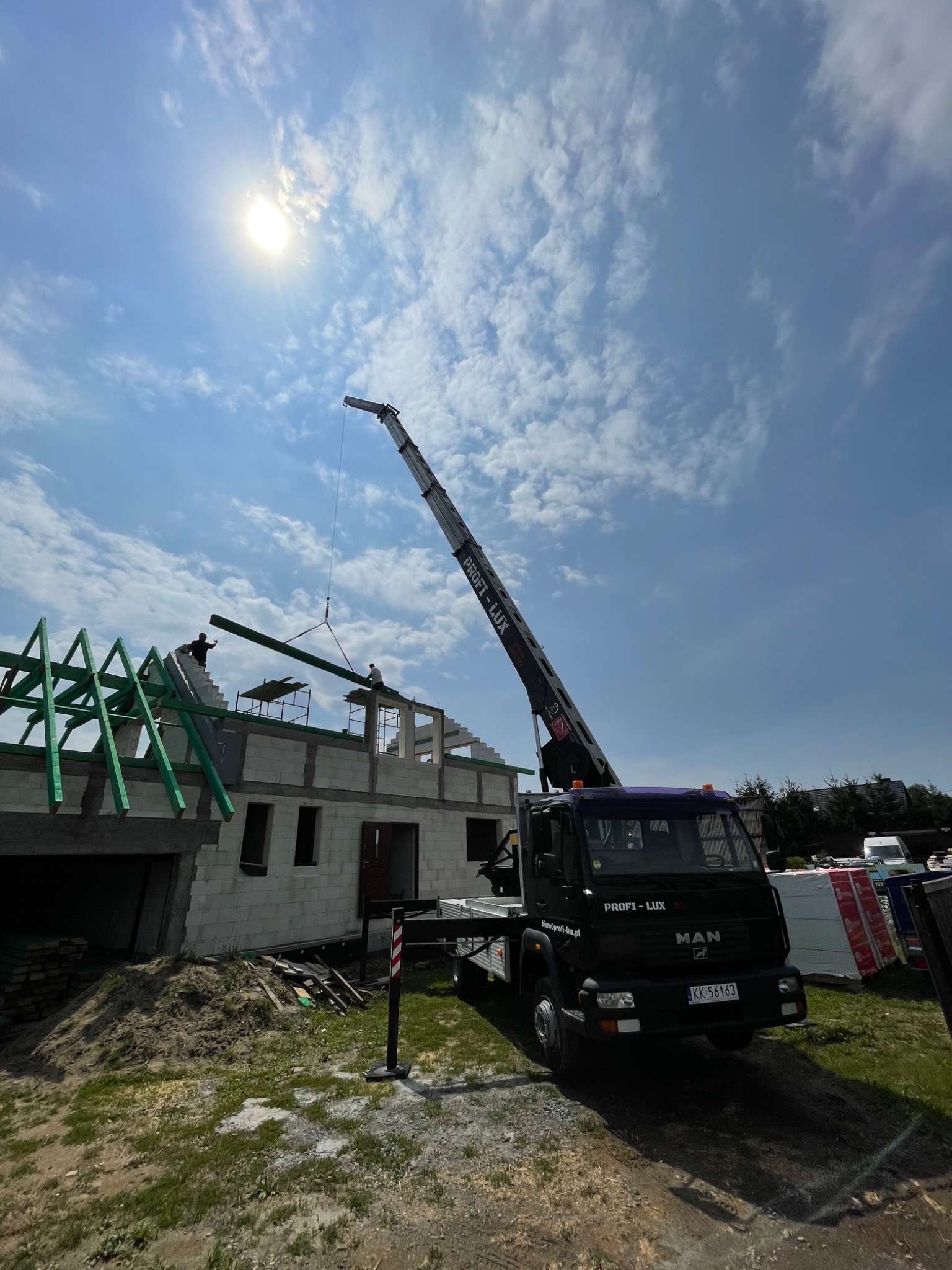 Czarny dźwig samochodowy marki MAN z wysięgnikiem podnosi element więźby dachowej na budowę domu z białego bloczka, na tle błękitnego nieba z chmurami i słońcem.
