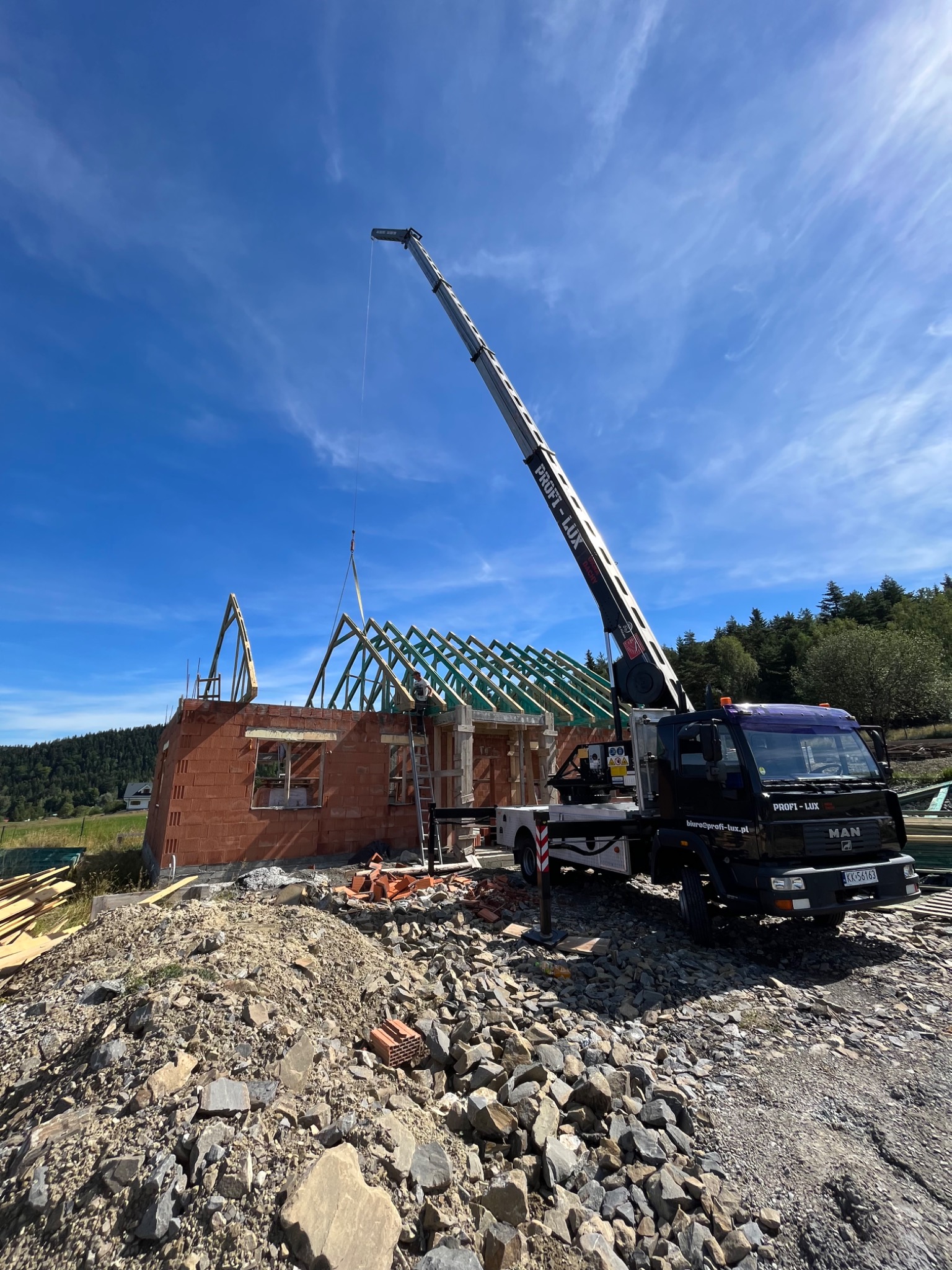 Czarny dźwig samochodowy MAN z wysięgnikiem PROFIT-LUX podnosi element więźby dachowej na budowie domu z czerwonej cegły, na tle błękitnego nieba i zielonego lasu.
