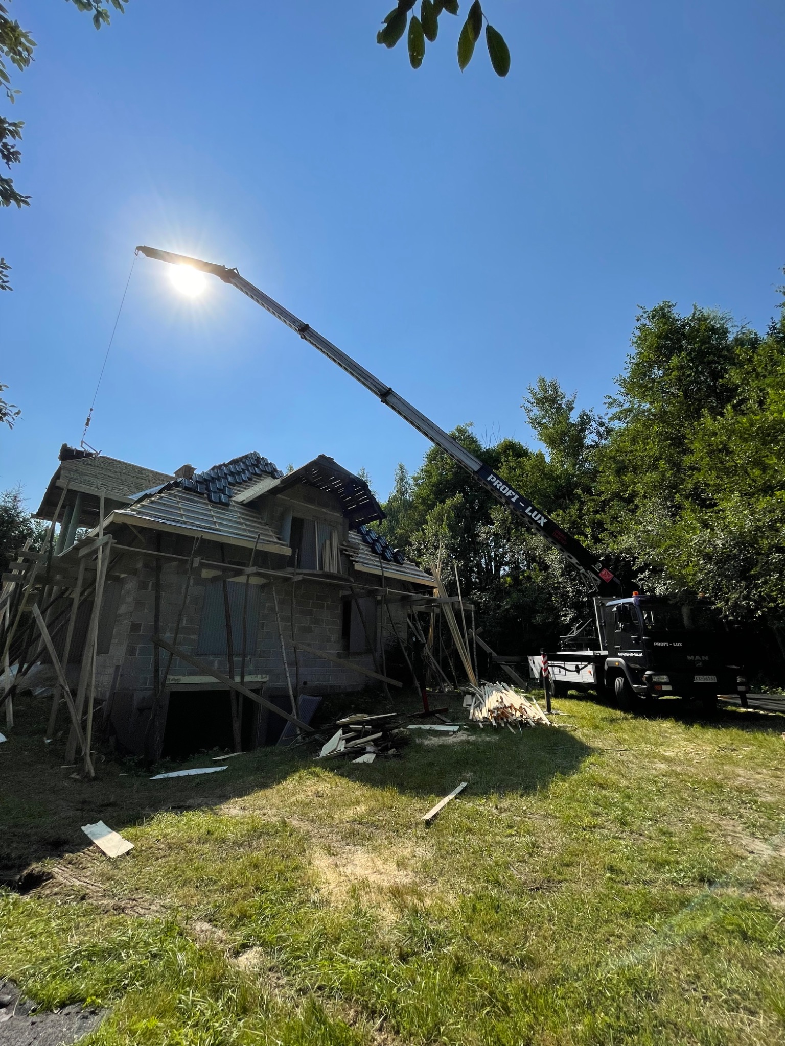 Montaż dachówki na budowie domu jednorodzinnego, widoczny dźwig z wysięgnikiem i robotnicy układający dachówkę, w tle las i błękitne niebo z promieniami słońca.