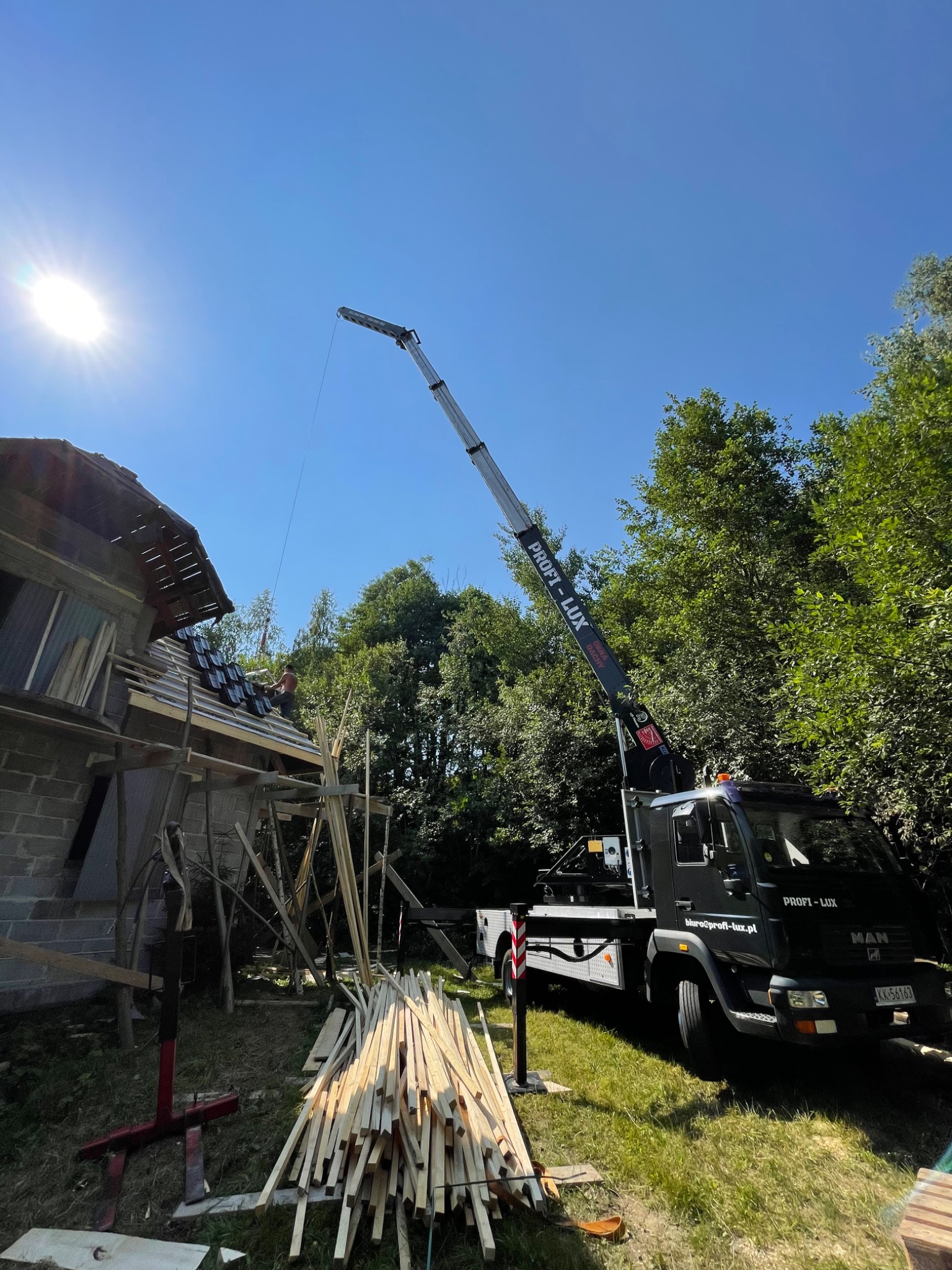 Czarny dźwig z wysięgnikiem podnosi elementy więźby dachowej na budowę domu jednorodzinnego w słoneczny dzień. Na pierwszym planie stos kantówek. Na dachu pracownik w trakcie układania dachówki.