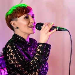 Kobieta z krótkimi rudymi włosami z zielonym podświetleniem, śpiewa do mikrofonu, ubrana w czarną koronkową bluzkę, z kolczykiem w kształcie kwadratu.
