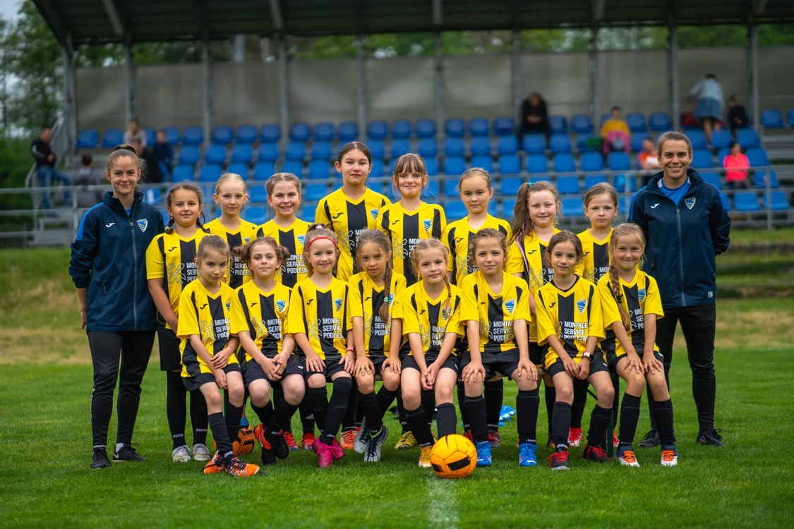 Drużyna młodych piłkarek w żółto-czarnych strojach pozuje do zdjęcia z trenerkami na tle trybun.