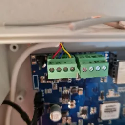 Detale podłączenia przewodów (czerwony, niebieski, żółty) do zielonych złączek na płytce alarmowej, widoczne śrubki mocujące przewody.