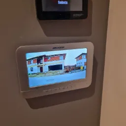 Ekran wideodomofonu Hikvision zamontowany na ścianie, wyświetlający obraz z kamery zewnętrznej przedstawiający nowo budowany dom w słoneczny dzień, z widocznym datownikiem i godziną. Dodatkowo...