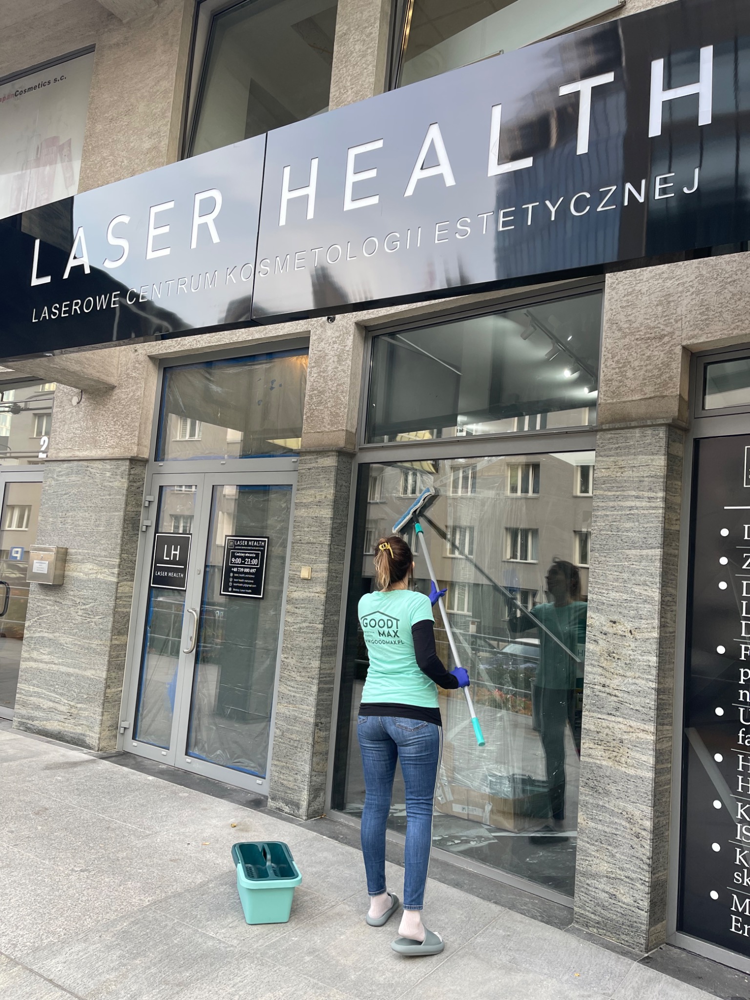 Kobieta w turkusowej koszulce i jeansach myje okno lokalu Laser Health za pomocą myjki teleskopowej; wiaderko z płynem stoi obok.