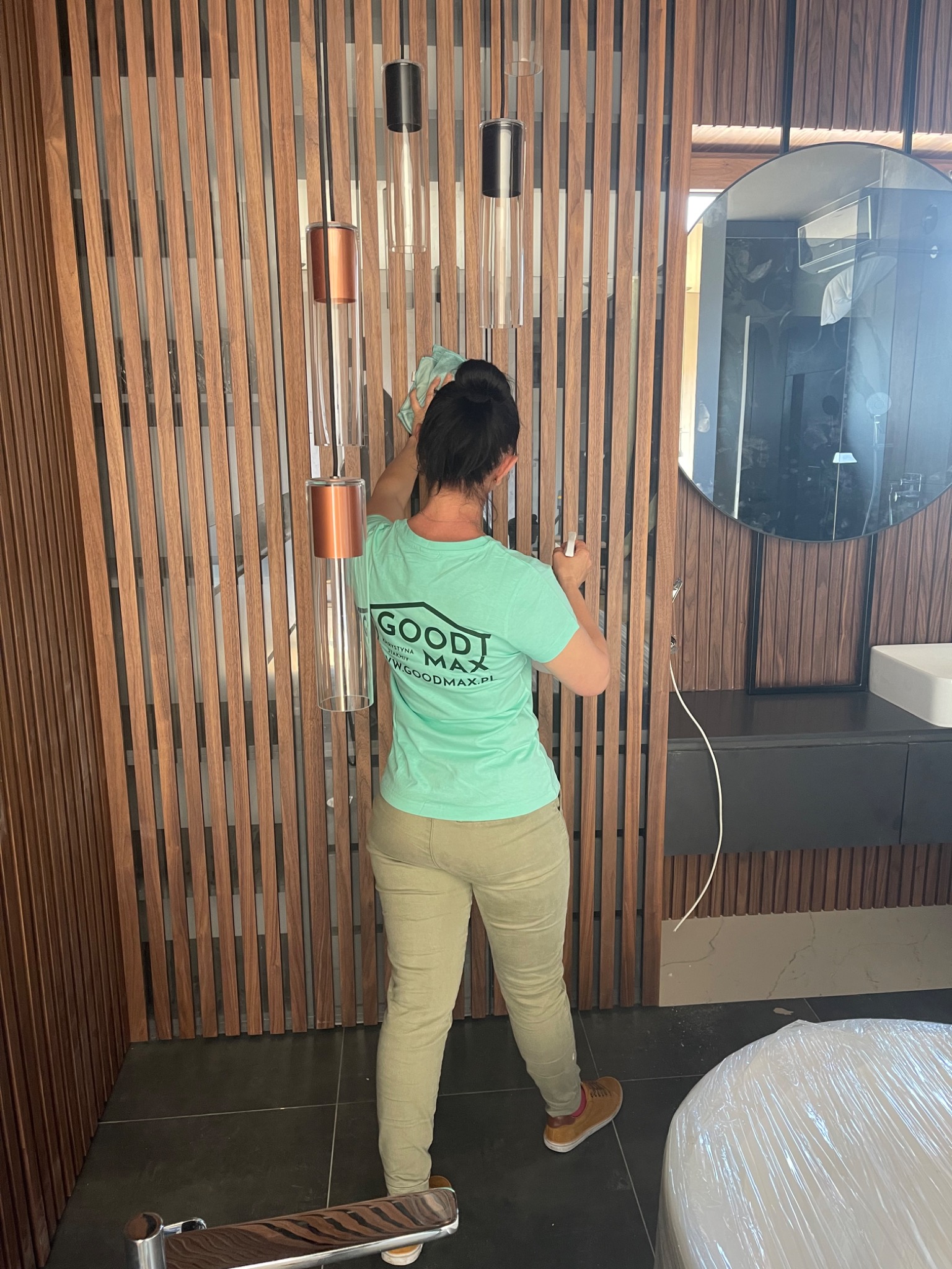 Pracownica firmy sprzątającej w zielonej koszulce z logo czyści szklane elementy wiszących lamp na tle drewnianej ściany w łazience z okrągłym lustrem.