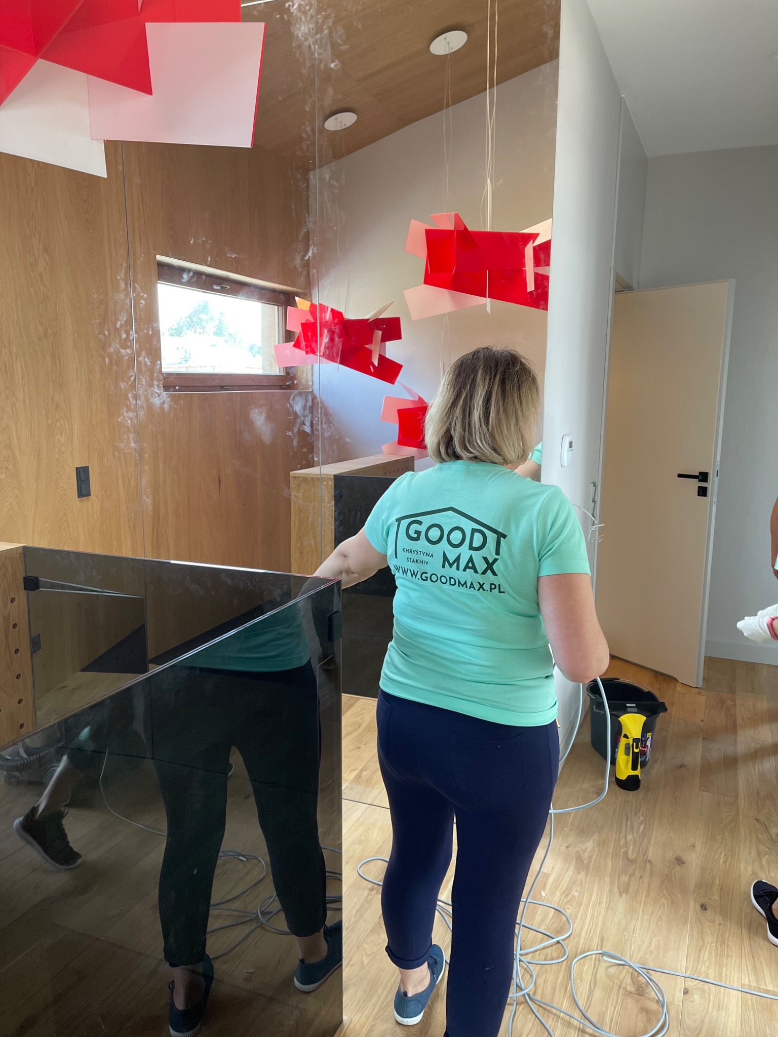 Pracownica firmy sprzątającej, w turkusowej koszulce z logo Goodt Max, czyści szklaną ścianę z pozostałości po remoncie, widoczne czerwone elementy dekoracyjne na suficie i ścianach.