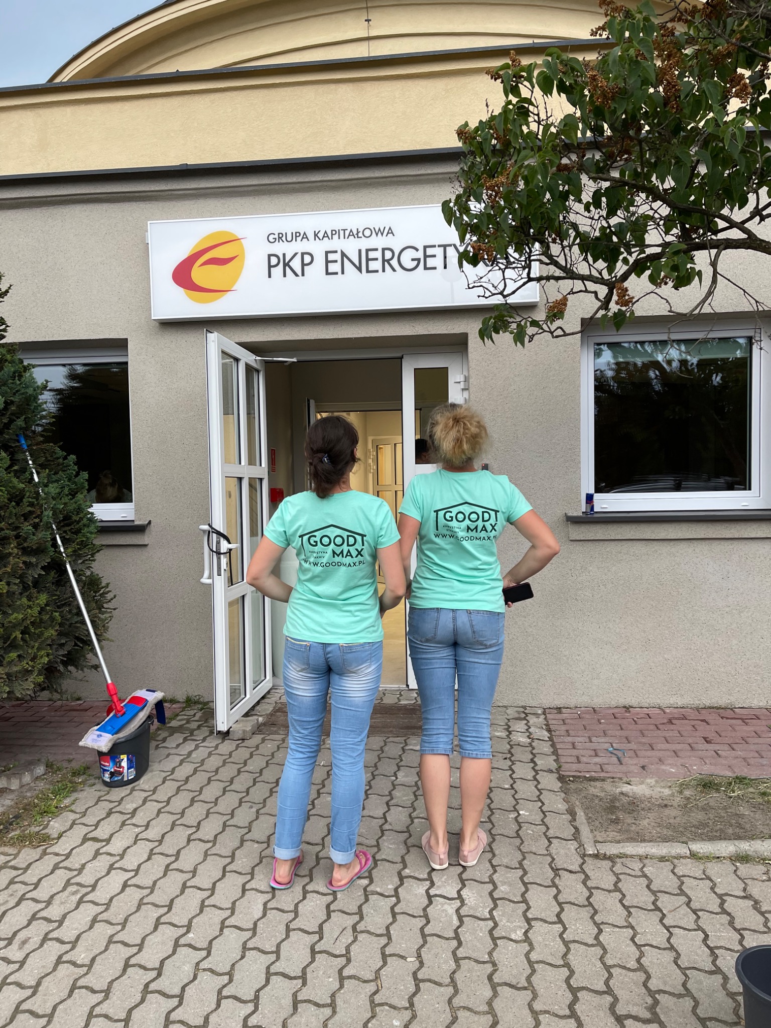 Dwie kobiety w jeansach i turkusowych koszulkach firmowych stoją przed budynkiem PKP Energetyka, obok widoczne wiadro i mop do podłogi.