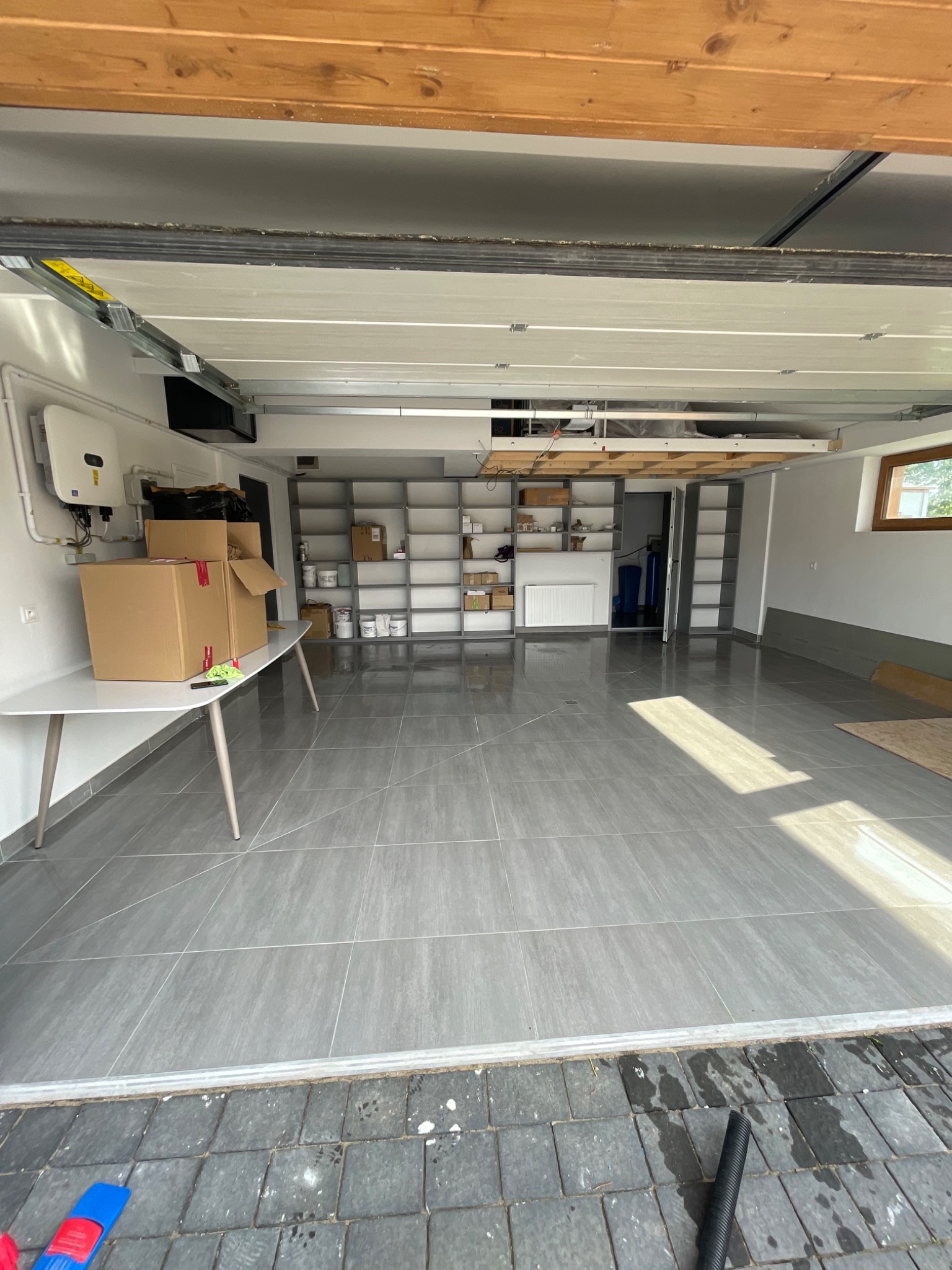 Wnętrze garażu w Warszawie po opróżnieniu, z szarymi płytkami na podłodze, białymi ścianami i regałami, oraz kartonami na stole po lewej stronie.